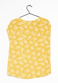 Gul blommig blus med vita blommor, korta ärmar, rund halsringning och rund nederkant. Lätt material med en slät textur.