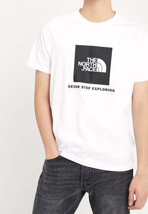 Osoba nosząca białą koszulkę z czarnym kwadratowym logo i tekstem "The North Face" oraz "Never Stop Exploring" w czarnych dżinsach.