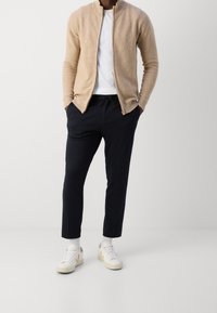 Beżowy kardigan na zamek, pod spodem biała koszulka, granatowe spodnie z troczkiem oraz białe trampki z beżowymi detalami. Materiał o fakturze, dopasowany krój.