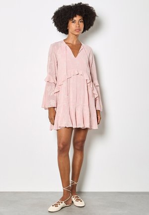 Donna con capelli ricci che indossa un mini abito rosa chiaro a maniche lunghe con volant e scarpe basse bianche con lacci, in piedi contro un muro semplice.