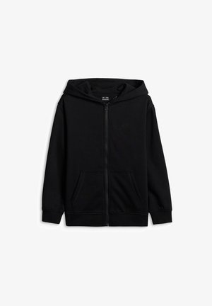 Garçon aux cheveux bouclés portant un sweat à capuche noir zippé, un t-shirt bleu, un pantalon noir, des chaussettes blanches, des baskets noir et blanc, et un bandeau noir à motifs, souriant.