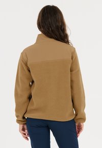 Vaaleanruskea fleece-takki, jossa on sileä, yhtenäinen yläosa. Pehmeä, teksturoidumpi alaosa, korkea kaulus ja pitkät hihat. Ei näkyviä kiinnityksiä.