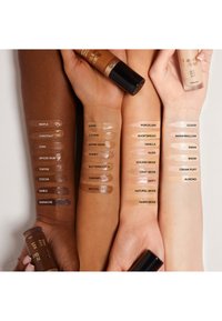 Trois bras présentent différents échantillons de fond de teint dans des tons de peau variés. Des nuances de marron foncé à beige clair sont étiquetées avec des noms descriptifs.
