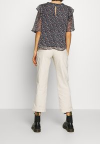 Blouse transparente à motifs floraux avec manches courtes à volants, détail dos avec ouverture en trou de serrure. Associée à un pantalon clair, légèrement ample, et des bottes noires.