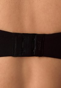 Čierny pás podprsenky s nastaviteľnými háčikmi na hladkom, svetlom povrchu kože. Obsahuje mäkký elastický materiál a šev pozdĺž okraja.