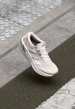 Chaussure de course Adidas rose clair avec des bandes blanches, flottant au-dessus d'un trottoir texturé près d'un bord et d'un pavage tactile.