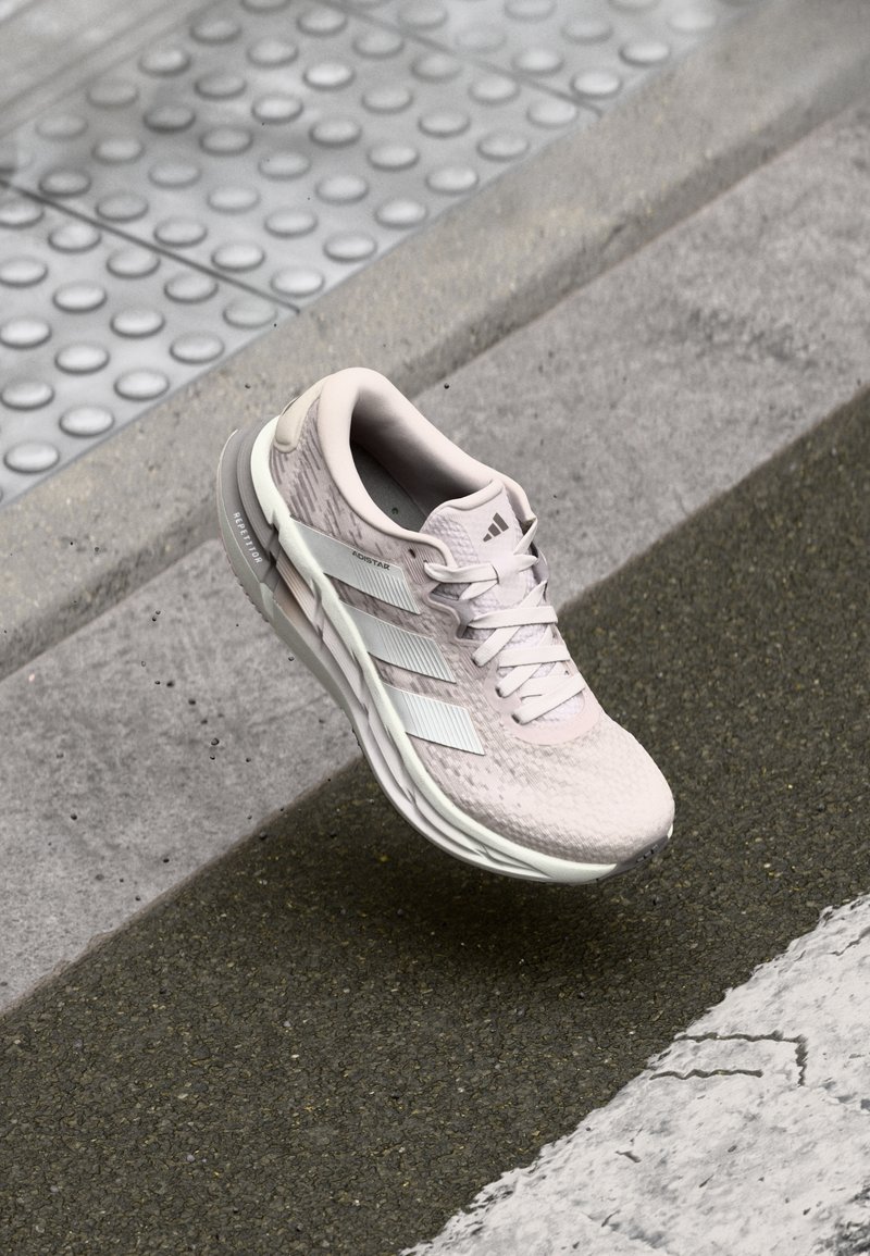 Chaussure de course Adidas rose clair avec des bandes blanches, flottant au-dessus d'un trottoir texturé près d'un bord et d'un pavage tactile.