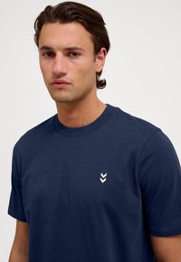 Mörkblå bomullströja med kort ärm, med en liten vit chevronlogotyp på vänster bröst och en ribbad rund halsringning.