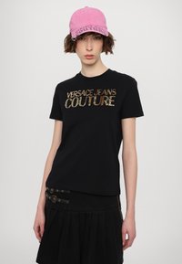 Črna majica s kratkimi rokavi z napisem "VERSACE JEANS COUTURE" iz kovinskih bleščic. Uporabljena z črno nabranem krilu z zlatim okovjem in roza kapo.