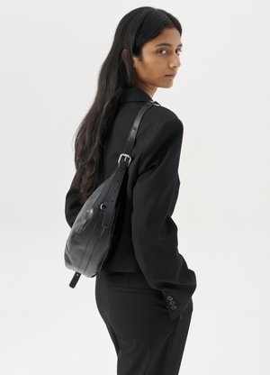 Femme avec de longs cheveux foncés portant un costume noir, portant un sac à épaule en cuir noir, regardant par-dessus son épaule contre un fond blanc.