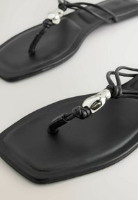 Sandales plates noires à bout carré, fines brides nouées et perles décoratives métalliques argentées sur fond clair.