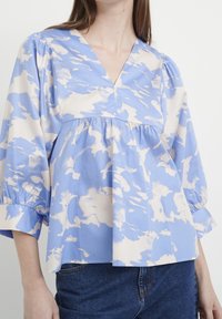 Modrá a krémová halenka s vzorem, s výstřihem do V, řasenými rukávy a volným střihem. Vyrobena z lehkého materiálu, s jemnou strukturou a splývavostí.