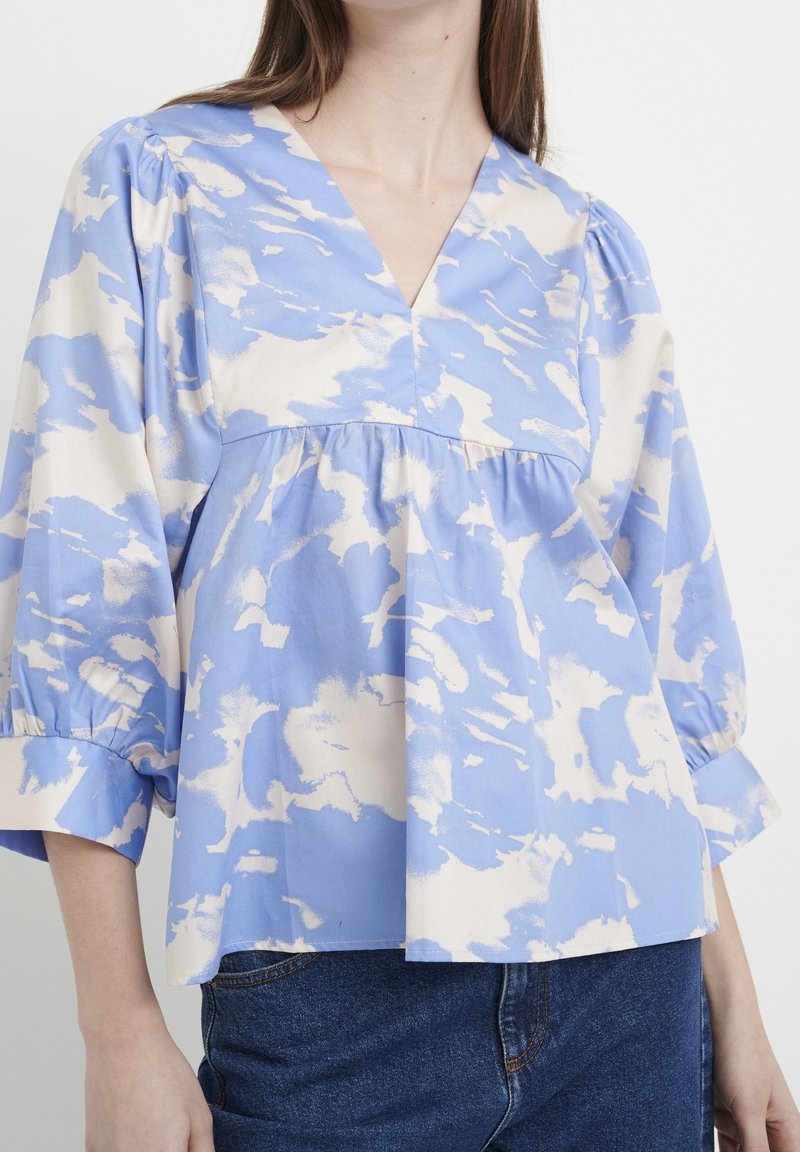 Modrá a krémová halenka s vzorem, s výstřihem do V, řasenými rukávy a volným střihem. Vyrobena z lehkého materiálu, s jemnou strukturou a splývavostí.