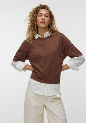 Kvinde med krøllet hår iført brun sweater over hvid skjorte og cremefarvede bukser står med hænderne på hofterne mod en ensfarvet baggrund.