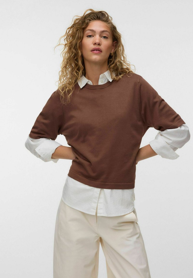 Femme aux cheveux bouclés portant un pull marron par-dessus une chemise blanche et un pantalon crème, debout les mains sur les hanches devant un fond uni.