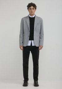 Grauer Blazer mit zwei Fronttaschen, getragen über einem schwarzen Pullover und einem weißen Hemd. Schwarze Slim-Fit-Hosen und schwarze Schuhe vervollständigen das Outfit.