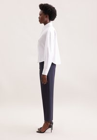 Camicia bianca con colletto e maniche lunghe, abbinata a pantaloni tapered blu. Sandali neri con tacco alto completano l'outfit. Tessuti lisci.