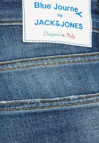 Tela de denim con un tono azul claro, que presenta una etiqueta que dice "Blue Journe by JACK & JONES", con costuras y detalles desgastados.
