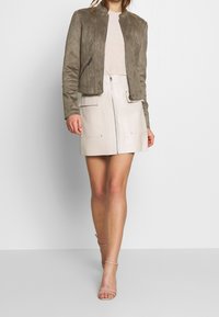 Chaqueta bomber de ante verde oliva, top beige ajustado, falda blanca de cuero con cremallera y bolsillos, y sandalias nude con tiras. Texturas suaves y colores audaces.