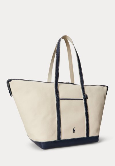 Stor beige lærredsnettaske med navyblå håndtag, kant og bund, med en lille broderet logo på frontlommen.