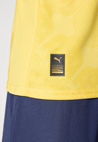 Gul sportsdrakt med en svart og gull Puma "Authentic Licensed Football"-etikett nær sømmen, båret over mørkeblå shorts.
