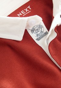 Red polo shirt with white collar. Features a button placket and side label with branding. Made of textured cotton fabric.