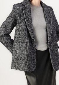 Schwarz-weiß kariertes Blazer mit Hahnentrittmuster, mit Reverskragen, Fronttaschen und tailliertem Schnitt, kombiniert mit einem grauen, strukturierten Oberteil.