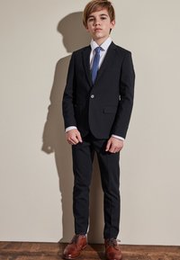 Czarny garnitur z dopasowaną marynarką i wąskimi spodniami, biała koszula i jasnoniebieski krawat. Brązowe eleganckie buty dopełniają strój.