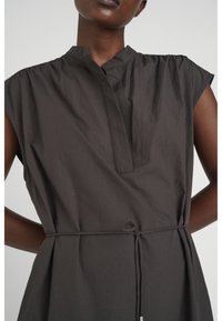 Mörk ärmlös klänning med mandarinkrage och smal midjeband bärs av en person med mörk hud mot en enkel ljus bakgrund.