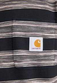 Stribet skjorte i sort og grå stof med en tekstureret følelse, der har et hvidt firkantet mærke med et gult Carhartt-logo og lomme detalje.