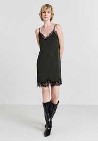 Donna che indossa un vestito a sottoveste nero senza maniche con pizzo e stivali in pelle neri al ginocchio, in piedi davanti a uno sfondo bianco semplice.