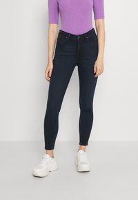 Pantalones vaquerosos ajustados de color azul oscuro con una textura suave, de talle alto y con bolsillos frontales, combinados con una parte superior ajustada de color morado claro y zapatillas blancas.