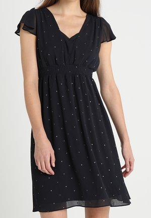 Femme portant une robe noire longueur genou avec de petits pois blancs, des manches papillon courtes et une taille cintrée.