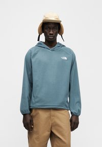 Φλις πουλόβερ σε τυρκουάζ με κουκούλα, με μικρό λευκό λογότυπο στο στήθος και ριμπ μανσέτες, συνδυασμένο με παντελόνι σε ταμπά και μπεζ καπέλο.