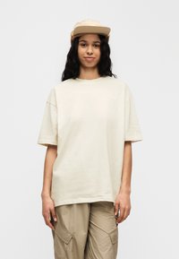 Μπεζ βαμβακερό φαρδύ t-shirt με κοντά μανίκια και στρογγυλή λαιμόκοψη. Συνδυασμένο με ανοιχτό χακί cargo παντελόνια και μπεζ καπέλο.