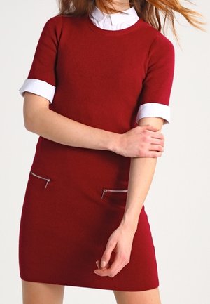 Femme portant une robe rouge à manches courtes avec un col et des poignets blancs, tenant son bras gauche avec sa main droite sur un fond uni.