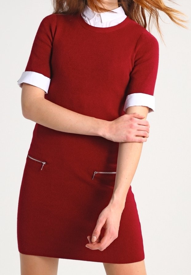 Femme portant une robe rouge à manches courtes avec un col et des poignets blancs, tenant son bras gauche avec sa main droite sur un fond uni.