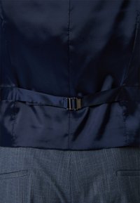 Marineblauw satijnen vest met een gladde textuur, voorzien van een riem aan de achterkant, gecombineerd met blauw geruite broek, wat het op maat gemaakte ontwerp benadrukt.