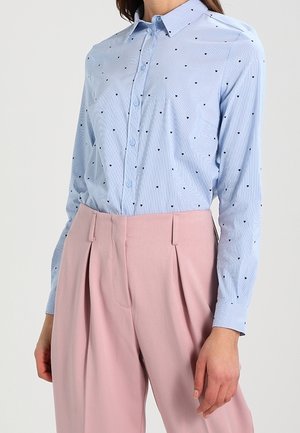 Femme portant une chemise bleu clair boutonnée avec de petits pois noirs et un pantalon plissé rose taille haute sur un fond uni.
