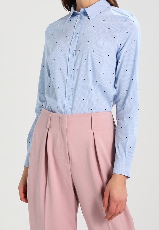 Femme portant une chemise bleu clair boutonnée avec de petits pois noirs et un pantalon plissé rose taille haute sur un fond uni.