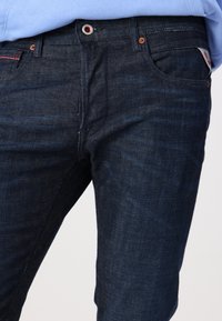 Gros plan sur un jean en denim bleu foncé porté avec une chemise bleu clair rentrée, montrant les poches avant, le bouton et les passants de ceinture.