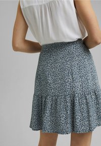 Femme portant une jupe bleue à fleurs avec un ourlet à volants et un chemisier blanc sans manches rentré, vue de dos.