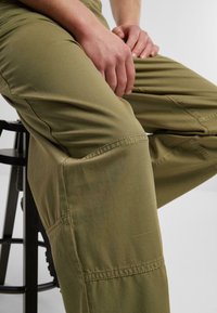 Pantalones cargo caqui con un ajuste relajado, que presentan detalles cosidos, bolsillos laterales y una tela de textura suave, apoyados en un taburete negro.