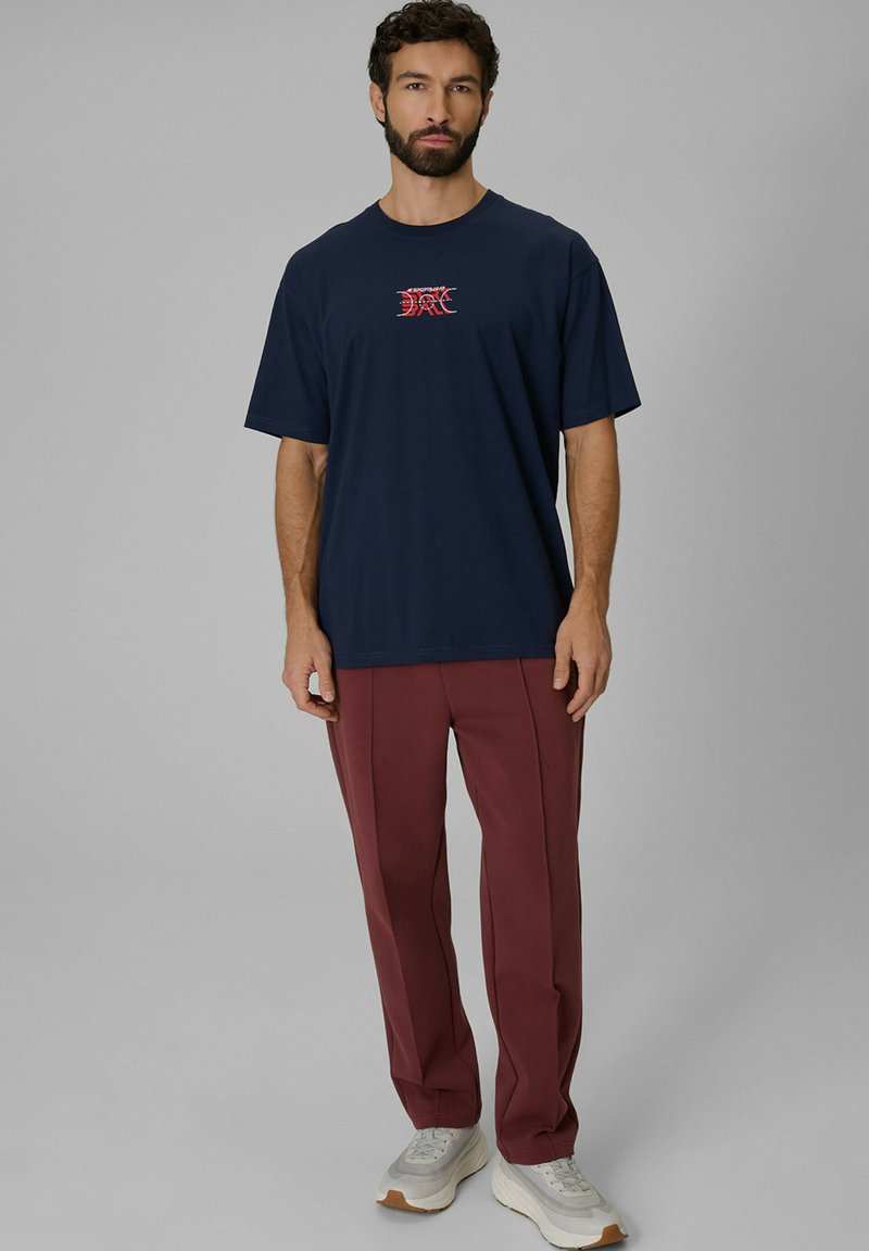Homme barbu portant un t-shirt bleu marine avec un graphisme rouge, un pantalon bordeaux et des baskets blanches, debout devant un fond gris.