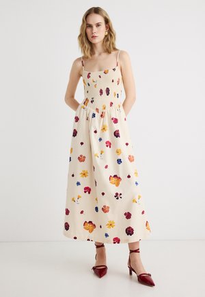 Γυναίκα με αμάνικο κρεμ φόρεμα με σμόκιν μπούστο και πολύχρωμο φλοράλ σχέδιο, συνδυασμένο με κόκκινα τακούνια με κορδόνια στους αστραγάλους, στέκεται μπροστά σε λευκό τοίχο.