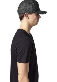 Sort kasket med et struktureret design og en flad skygge, lavet af tekstureret stof, båret af en person i en sort t-shirt, set fra siden.