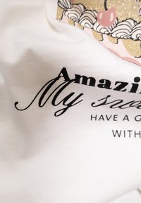 Camiseta de algodón blanca con texto en negro escrito "Increíble Mi..." y diseño de dibujos animados. Presenta acentos de purpurina rosa y dorada.