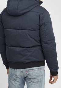 Granatowa kurtka puchowa z kapturem, pikowanym wzorem, elastycznymi mankietami i prążkowanym obszyciem, zestawiona z jasnoniebieskimi jeansami.