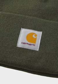 Green knitted beanie with a textured fabric and a square white tag featuring a yellow logo and red text.