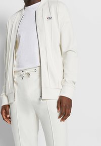 Witte jas met ritssluiting, geribbelde manchetten en verticale naadtoeslagen. Gecombineerd met bijpassende broek met een elastische tailleband en aantrekkoord.